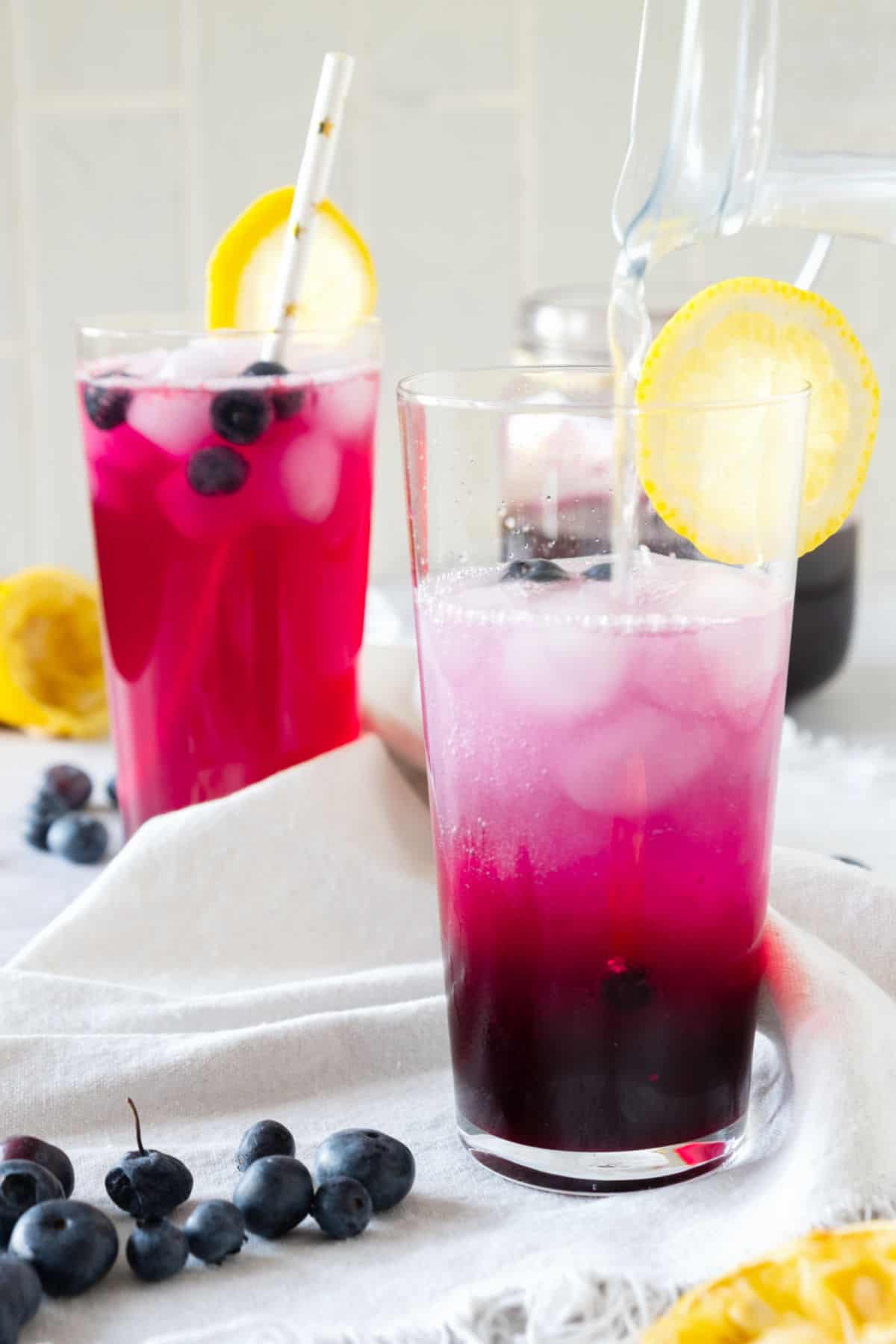 Pouring lemonade mix on tall glass with blueberry syrup, ice and vodka. White cloth as surface. Beige background.