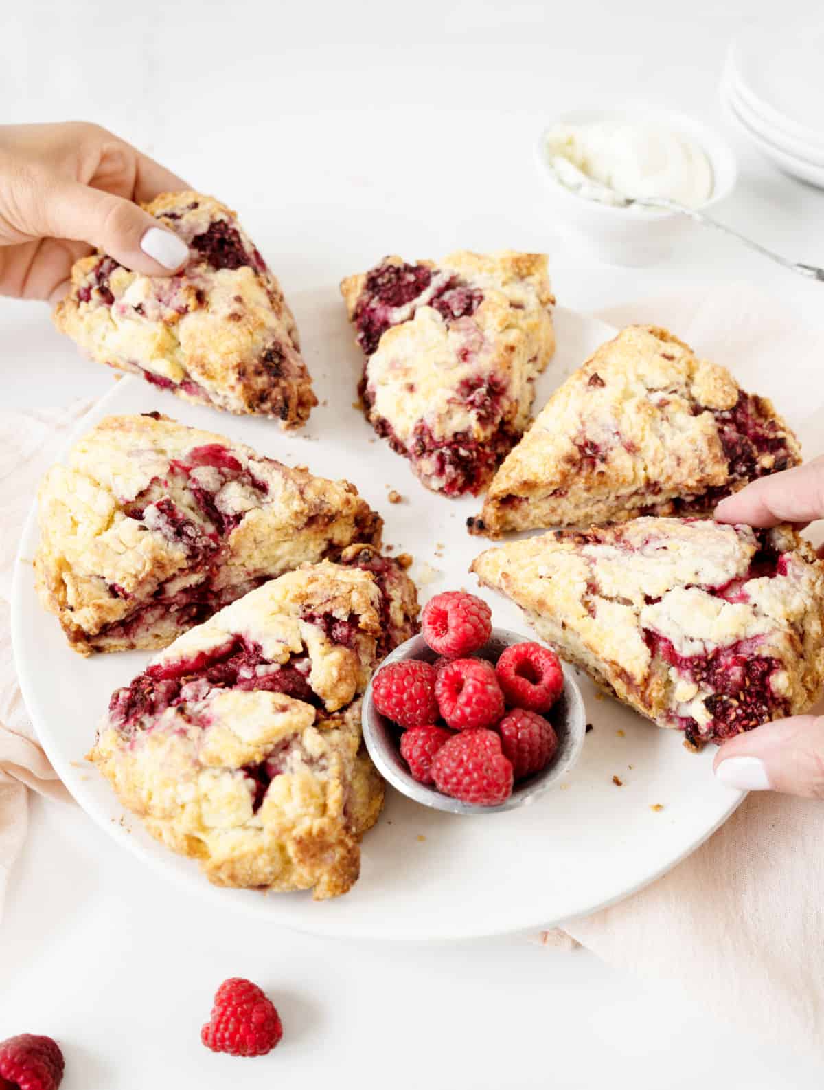 Hands lifting two raspberry lemon scones from a white plate. Pink white surface.