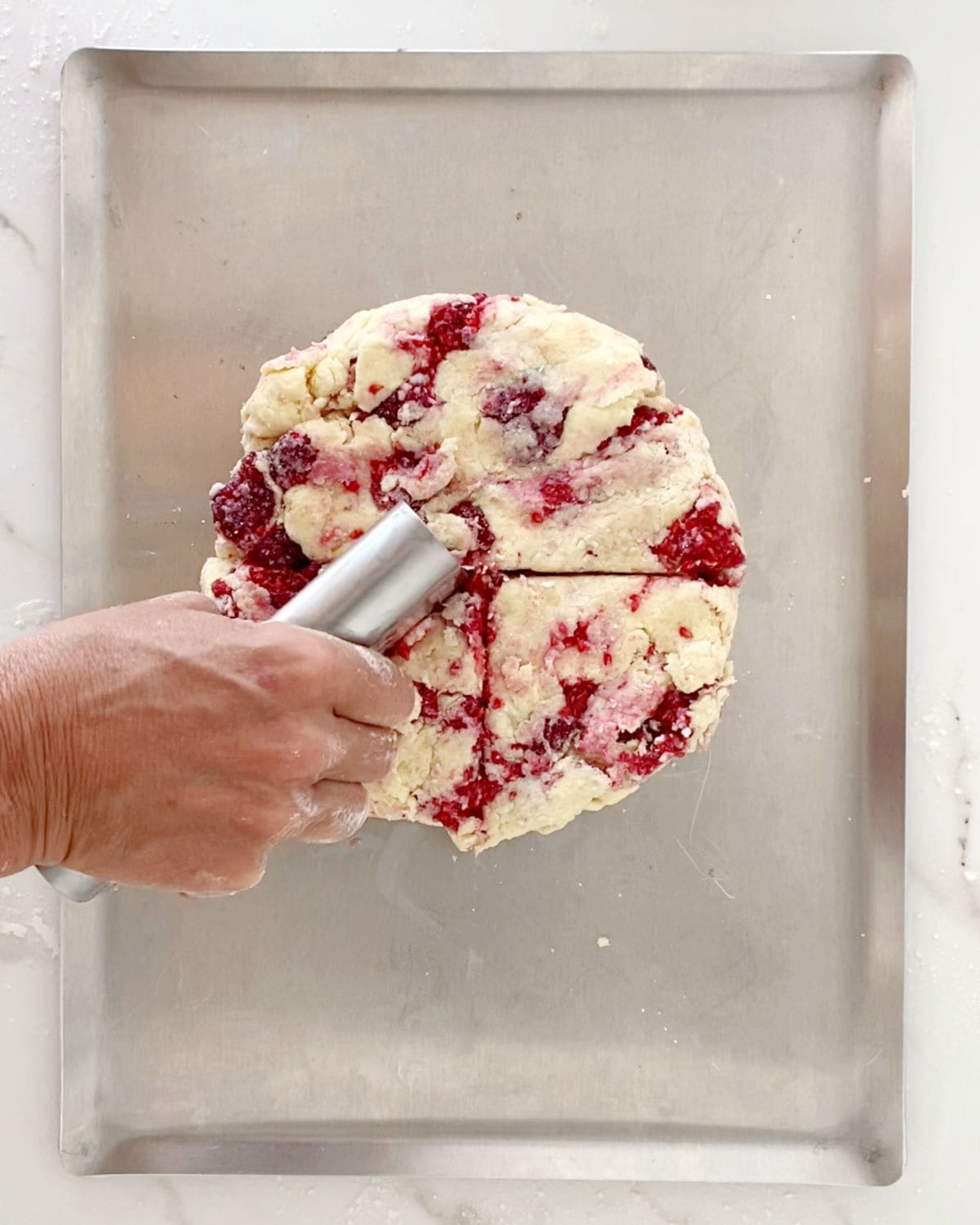 Cutting triangles of raspberry scones with a dough scraper on a metal baking sheet.