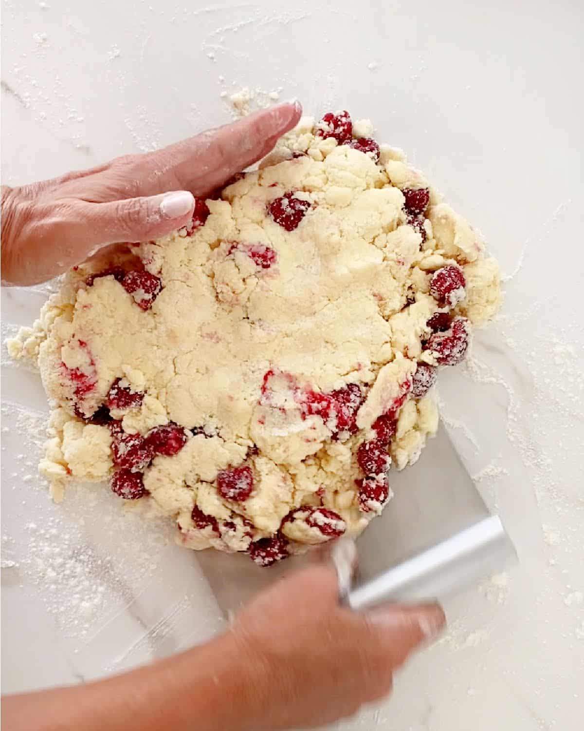 Forming raspberry scone dough disc with a dough scraper on a white marbled surface.