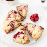Close up of five raspberry scones on a white plate. Small bowl of raspberries. White surface.