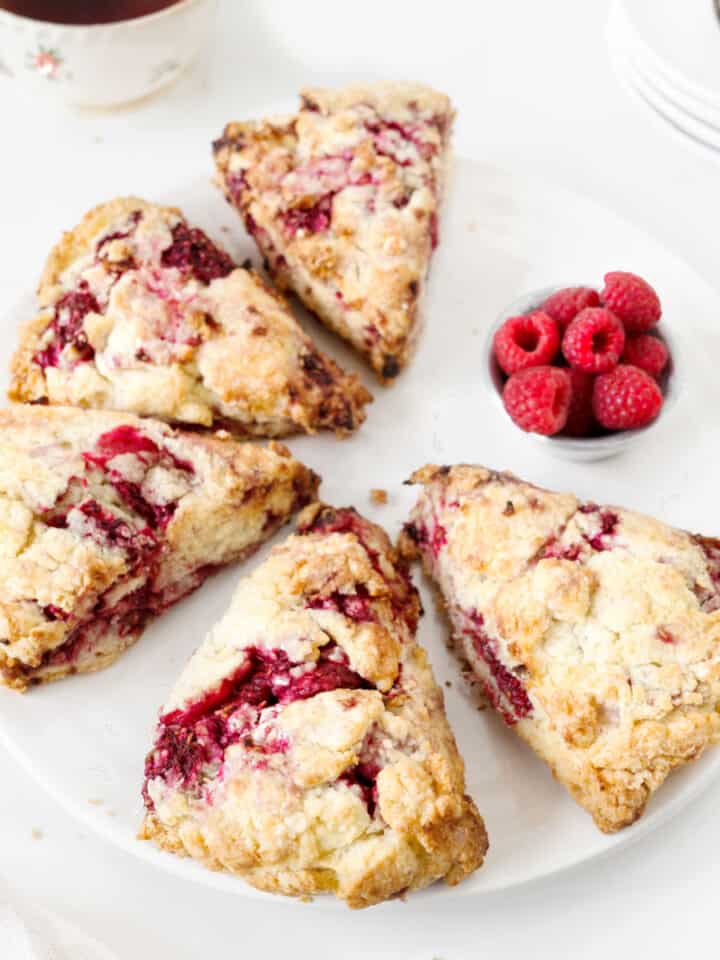 Close up of five raspberry scones on a white plate. Small bowl of raspberries. White surface.
