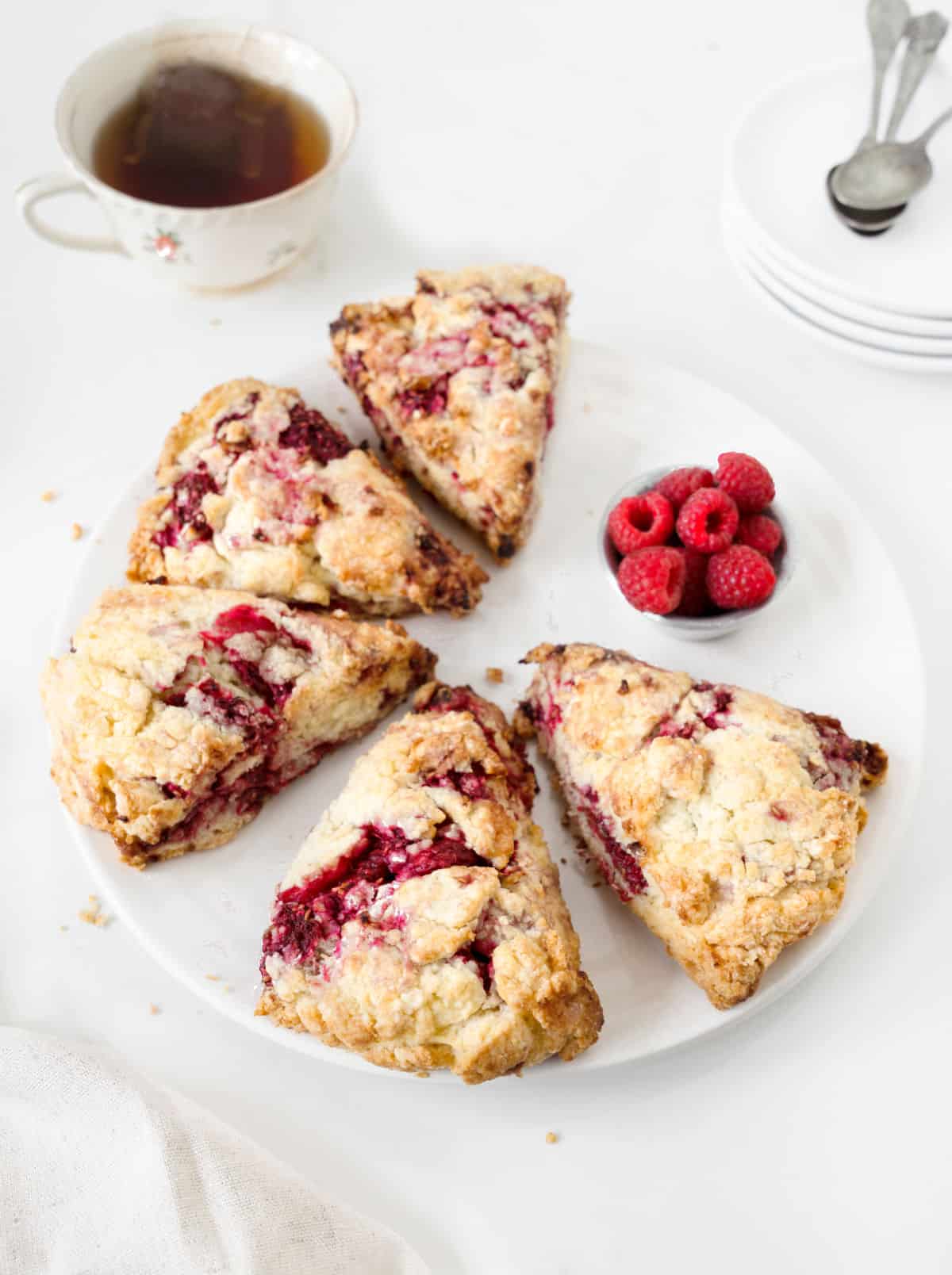 Several raspberry lemon scones on a white plate. Bowl of raspberries, cup of tea, white surface.