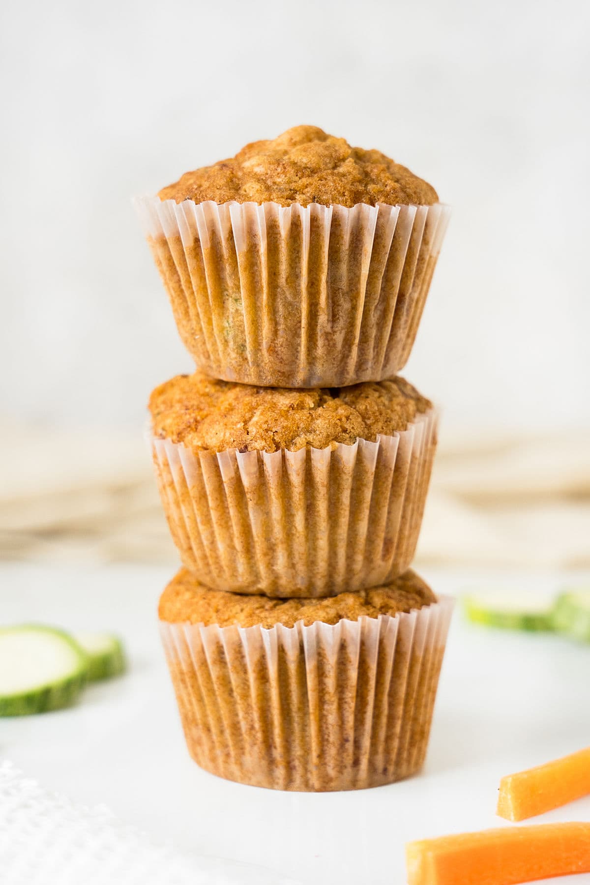 Three stacked carrot zucchini muffins in paper liners. White surface and background.