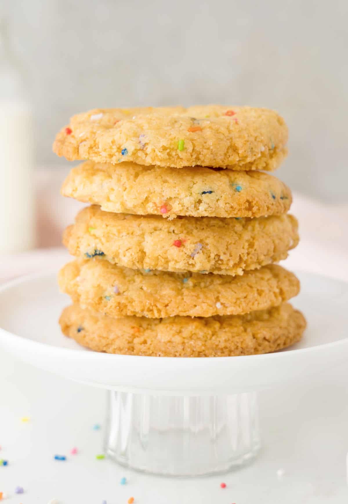Stack of funfetti cookies on a glass cake stand. White surface, grey background.