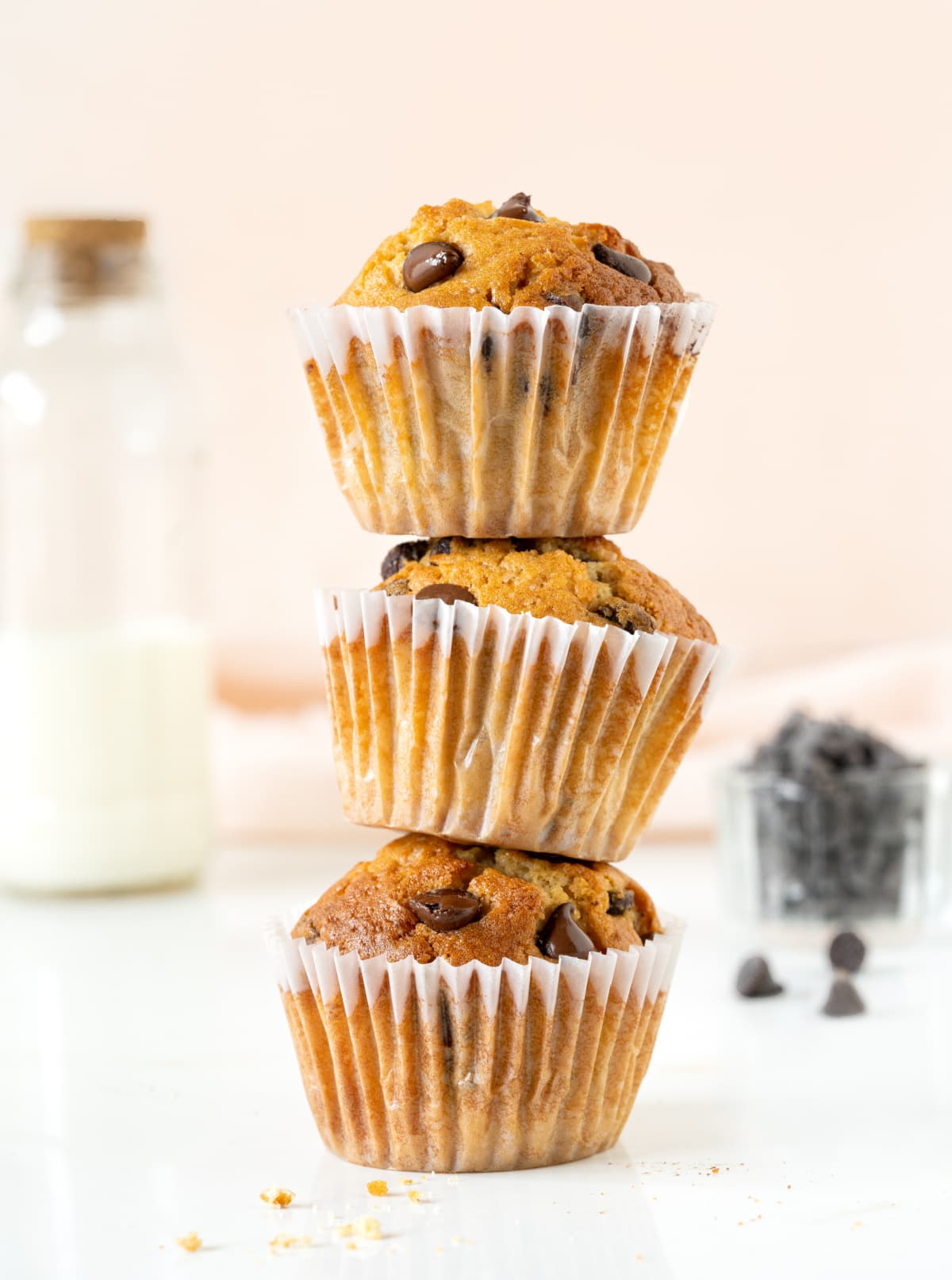 Stack of three chocolate chip muffins. White surface, pink background. Bowl of chips, milk bottle.