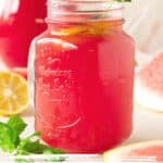 Pink and white text overlay on small jug of watermelon lemonade. Mint, lemon slices, pitcher; white surface, beige background.