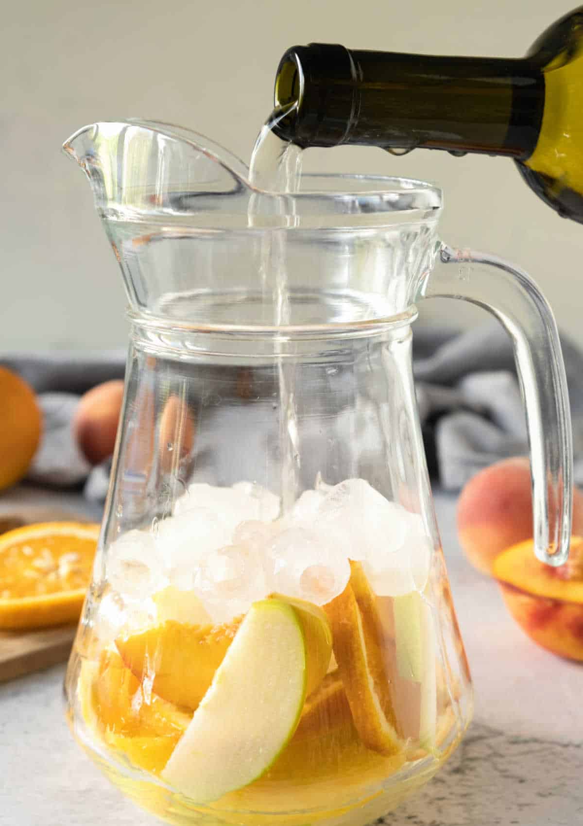 Pouring white wine from a bottle in a pitcher with ice and sliced fruit. Greyish background.