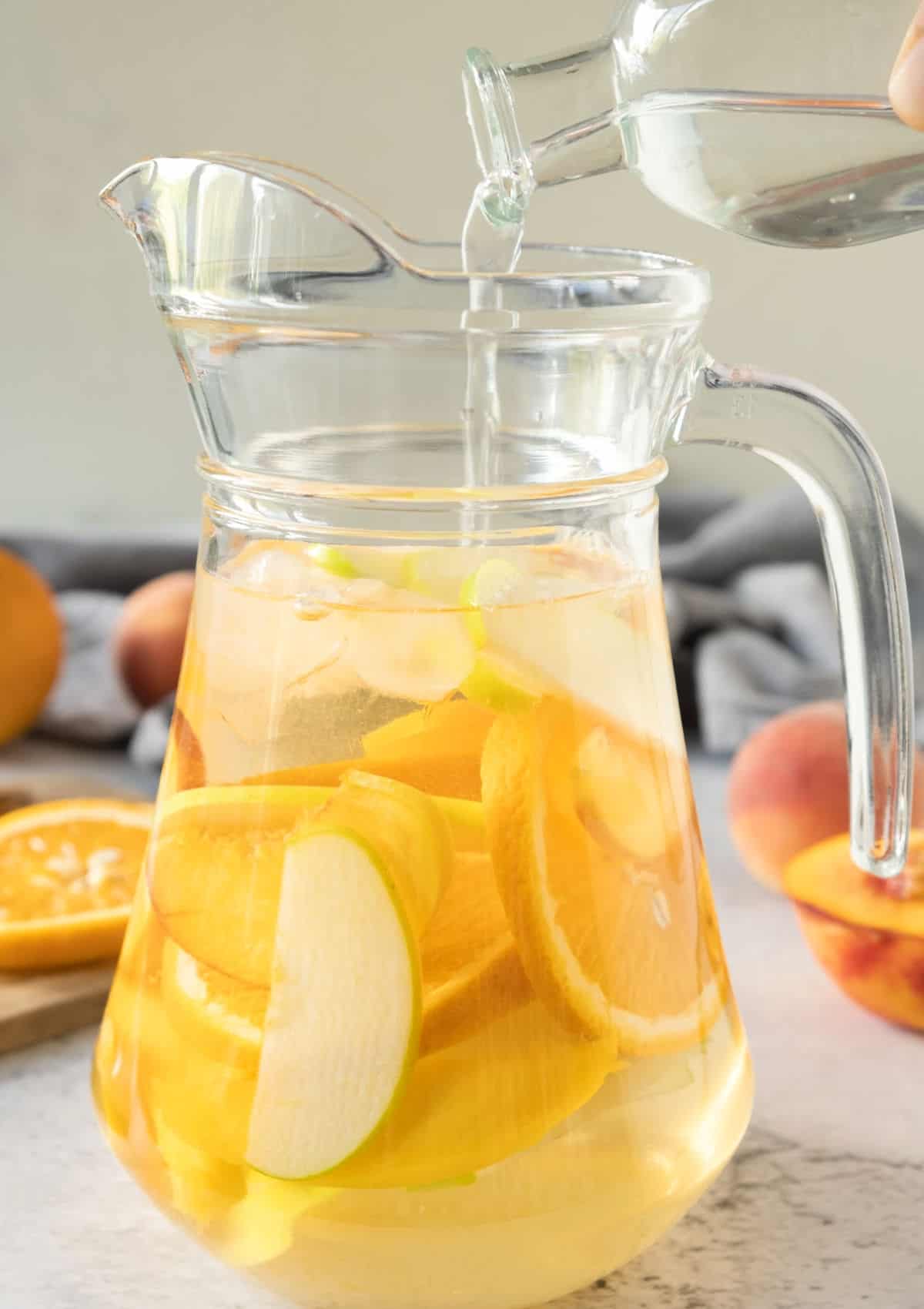 Sliced fruit and ice in a pitcher. Liquid being poured in. Grey surface and background.