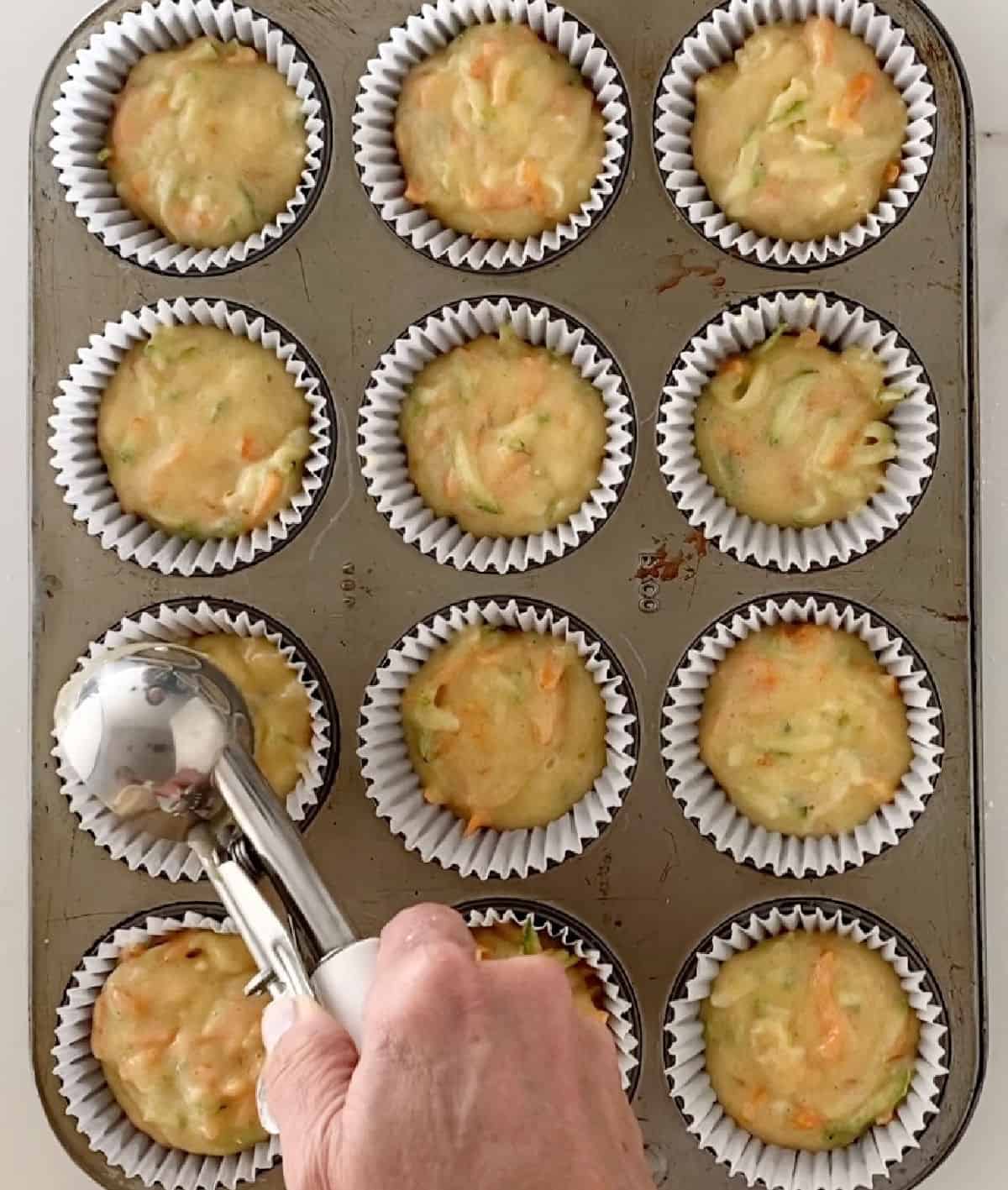 Scooping carrot zucchini muffin batter onto paper liner in a muffin pan.