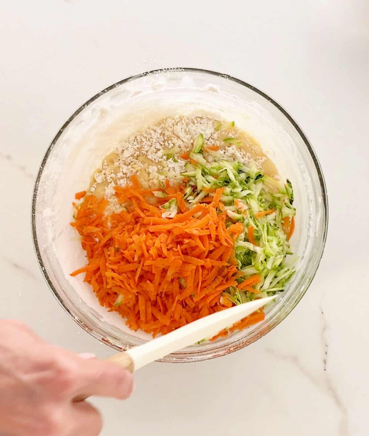 Grated zucchini and carrot added to muffin mixture in a glass bowl. White marbled surface. Hand holding a spatula.