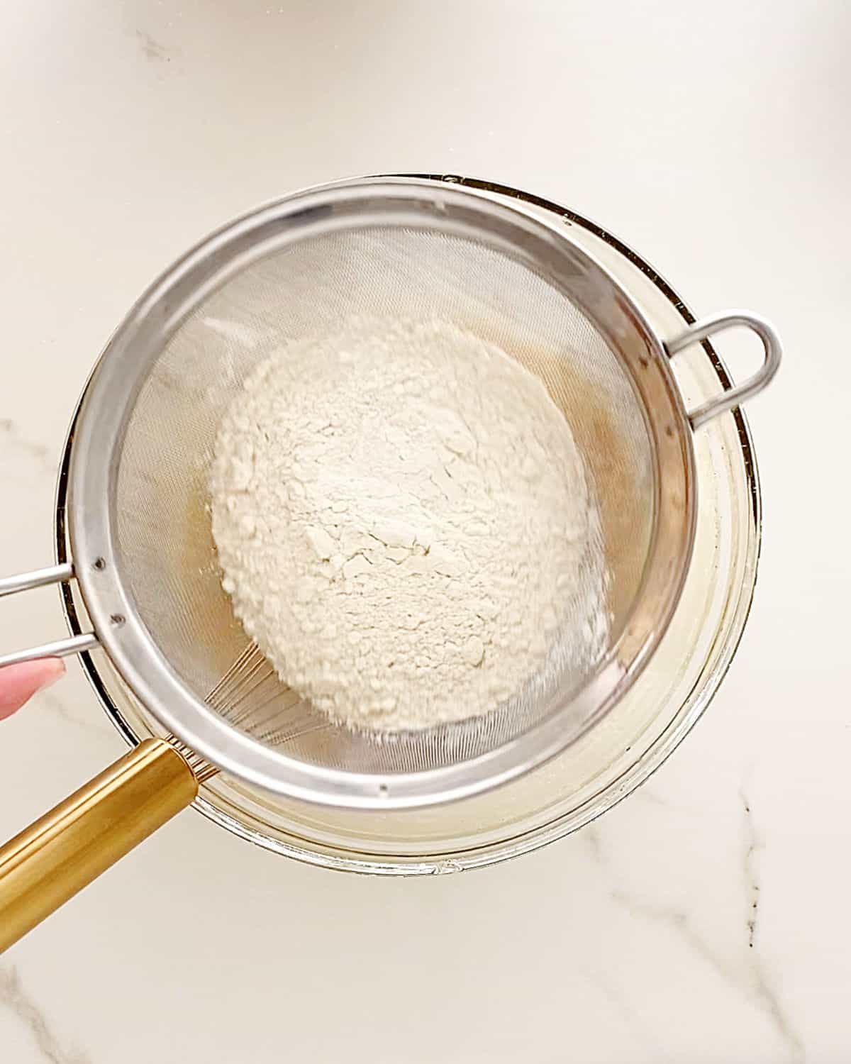 Sifting flour mixture over wet ingredients in a glass bowl. White surface. 