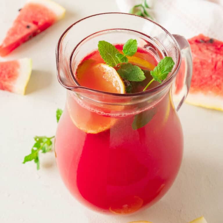 Pitcher of watermelon lemonade on an off-white surface, watermelon slices, mint leaves.