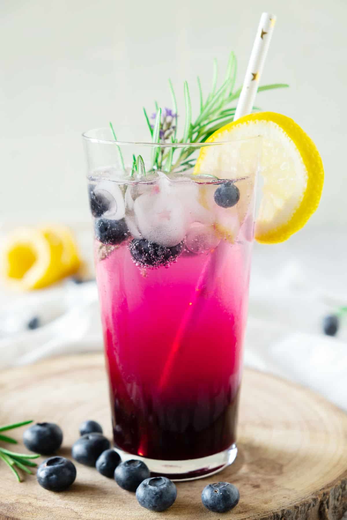 Single glass of blueberry vodka lemonade with rosemary sprig, lemon slice and ice. Wooden round, beige background.