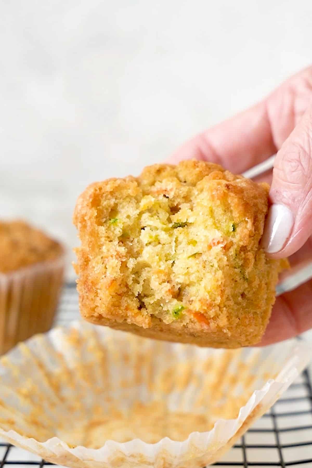 Holding a bitten zucchini carrot muffin over an opened paper liner. Greyish background.