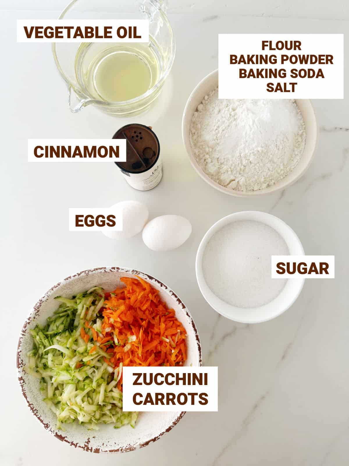 Bowls with ingredients for zucchini carrot muffins including oil, cinnamon, sugar, flour mixture. White marbled surface.