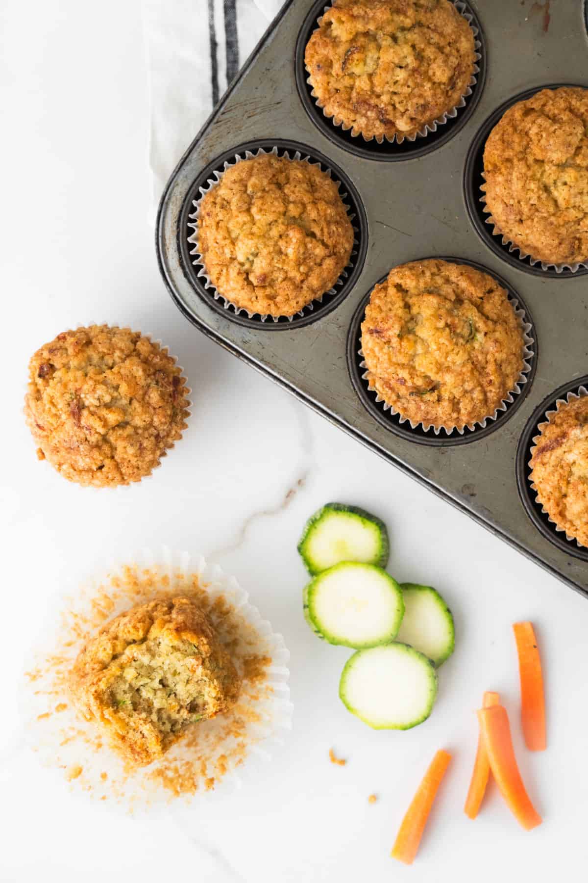 Top view of muffin pan with baked zucchini carrot muffins. White marble, zucchini and carrot slices, half a muffin.
