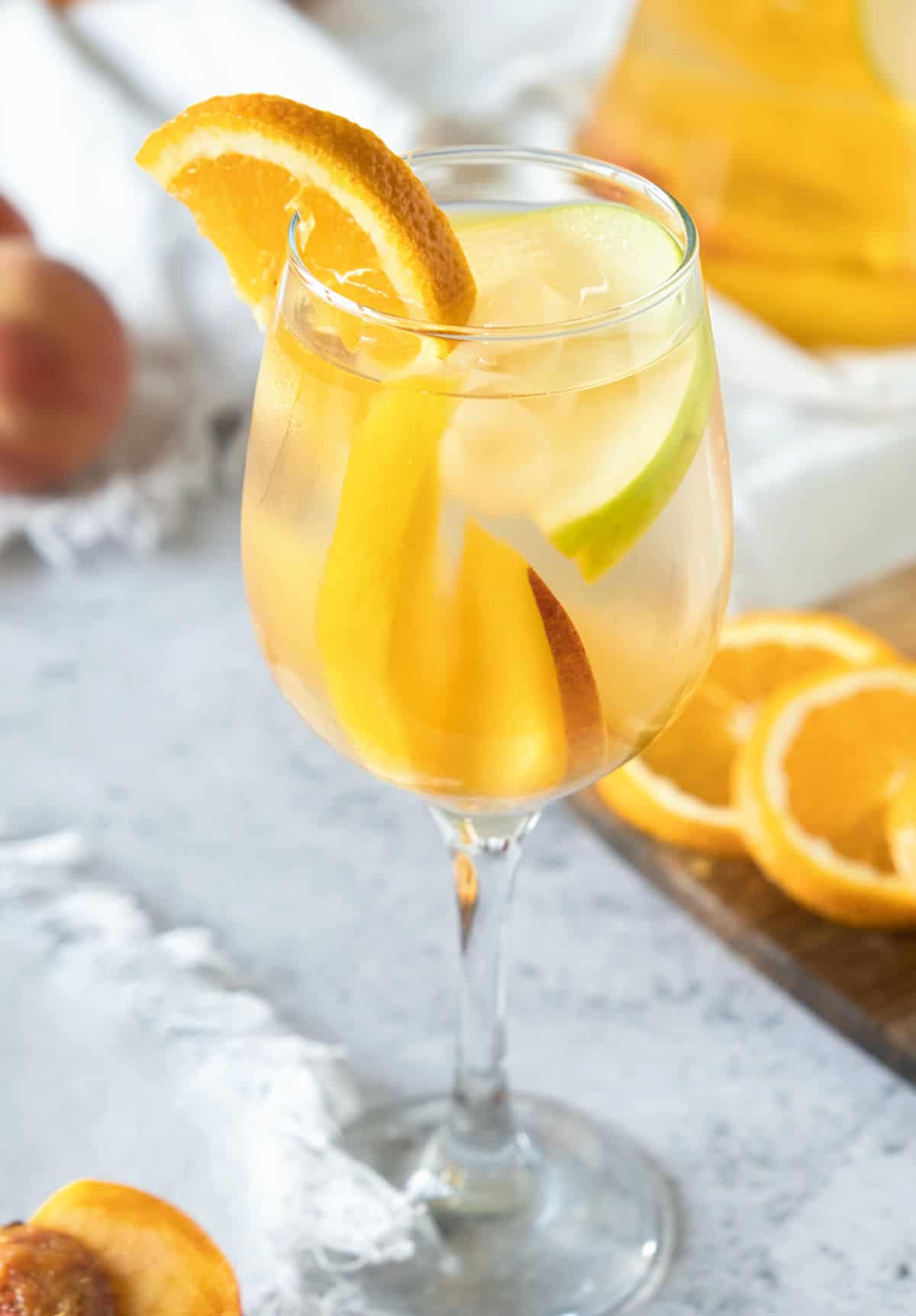 Stem glass with white wine sangria with orange and apple slices. Grey surface with more fruit. 