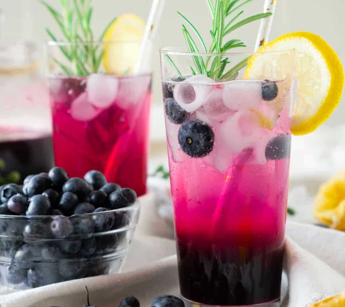 Bowl of blueberries and tall glasses of blueberry lemonade with rosemary sprigs and lemon slices.