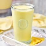 Tall glass of mango milkshake on a white plate. Yellow cloth on grey background.