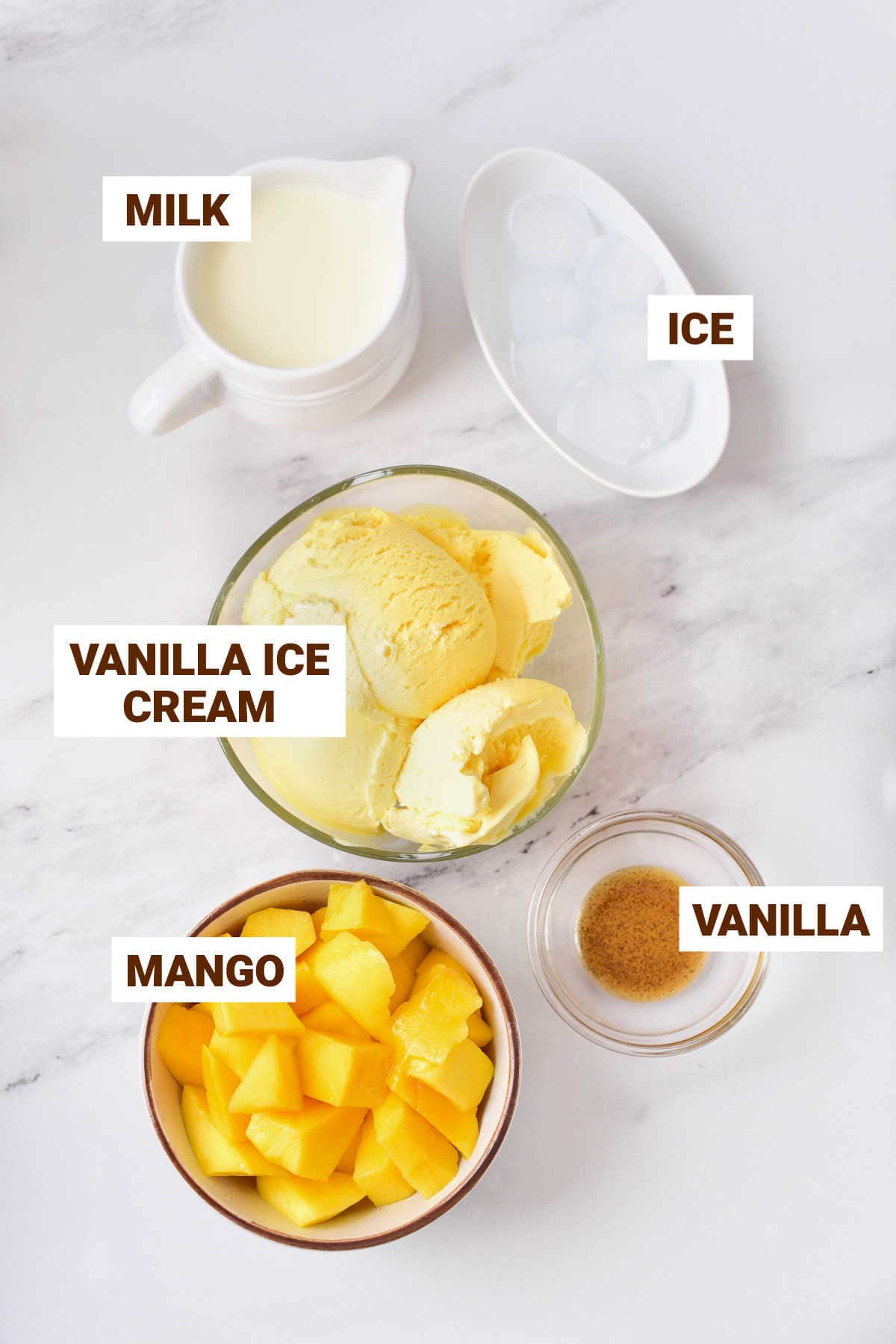 White marbled surface with bowl containing ingredients for mango milkshake including ice cream, milk, ice, vanilla.