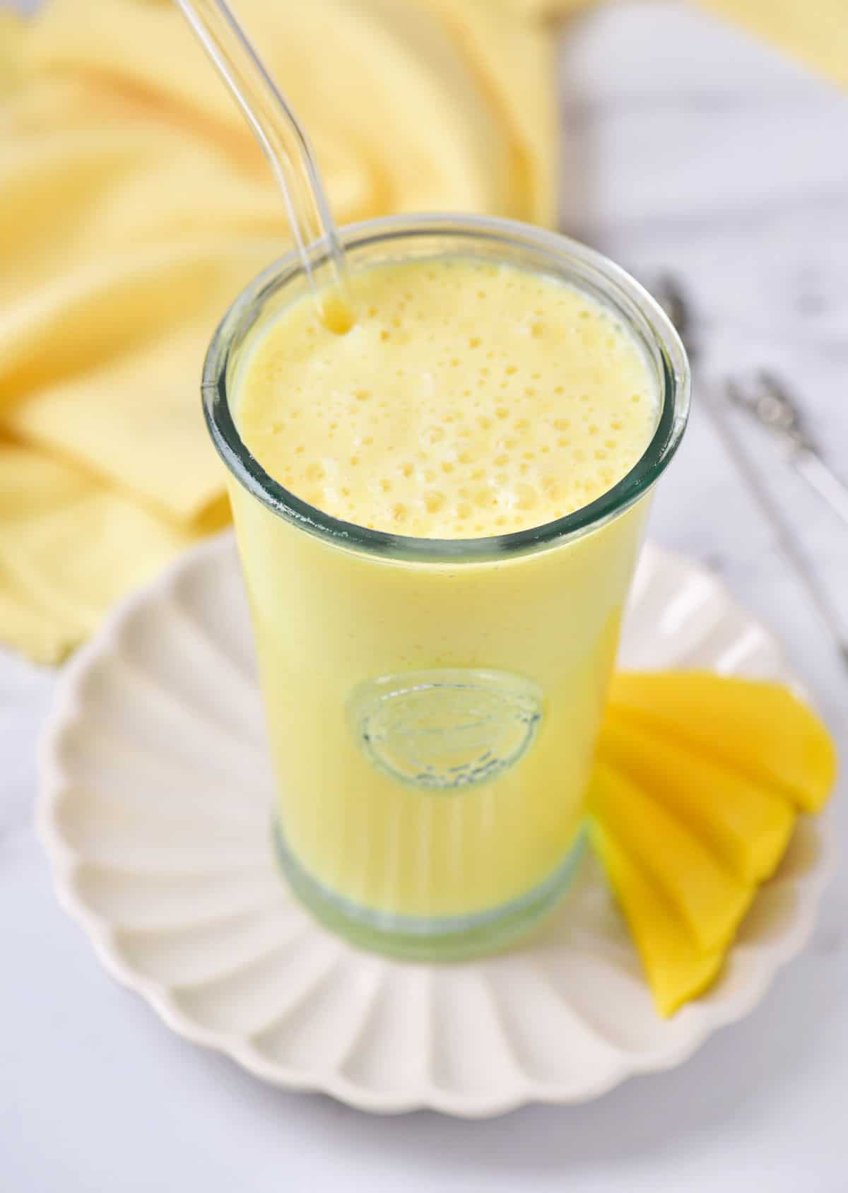 White plate with tall glass of mango milkshake. White marble surface and yellow cloth.