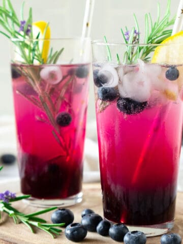 Two tall glasses of spiked blueberry lemonade with lemon slices and rosemary sprigs. Beige background.