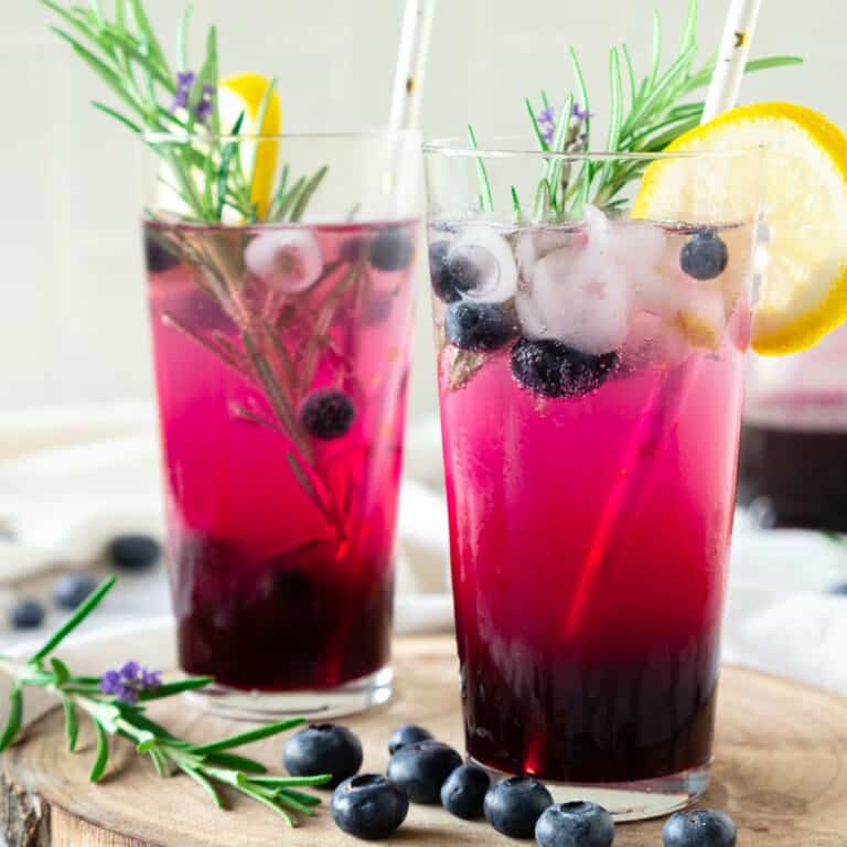 Two tall glasses of spiked blueberry lemonade with lemon slices and rosemary sprigs. Beige background.