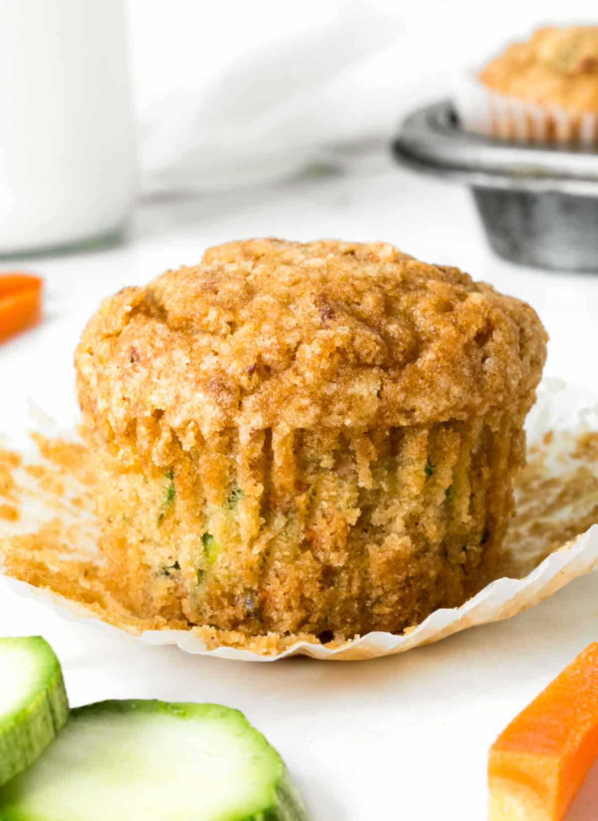 Opened paper liner with carrot zucchini muffin. White surface, zucchini and carrot pieces, muffin pan.