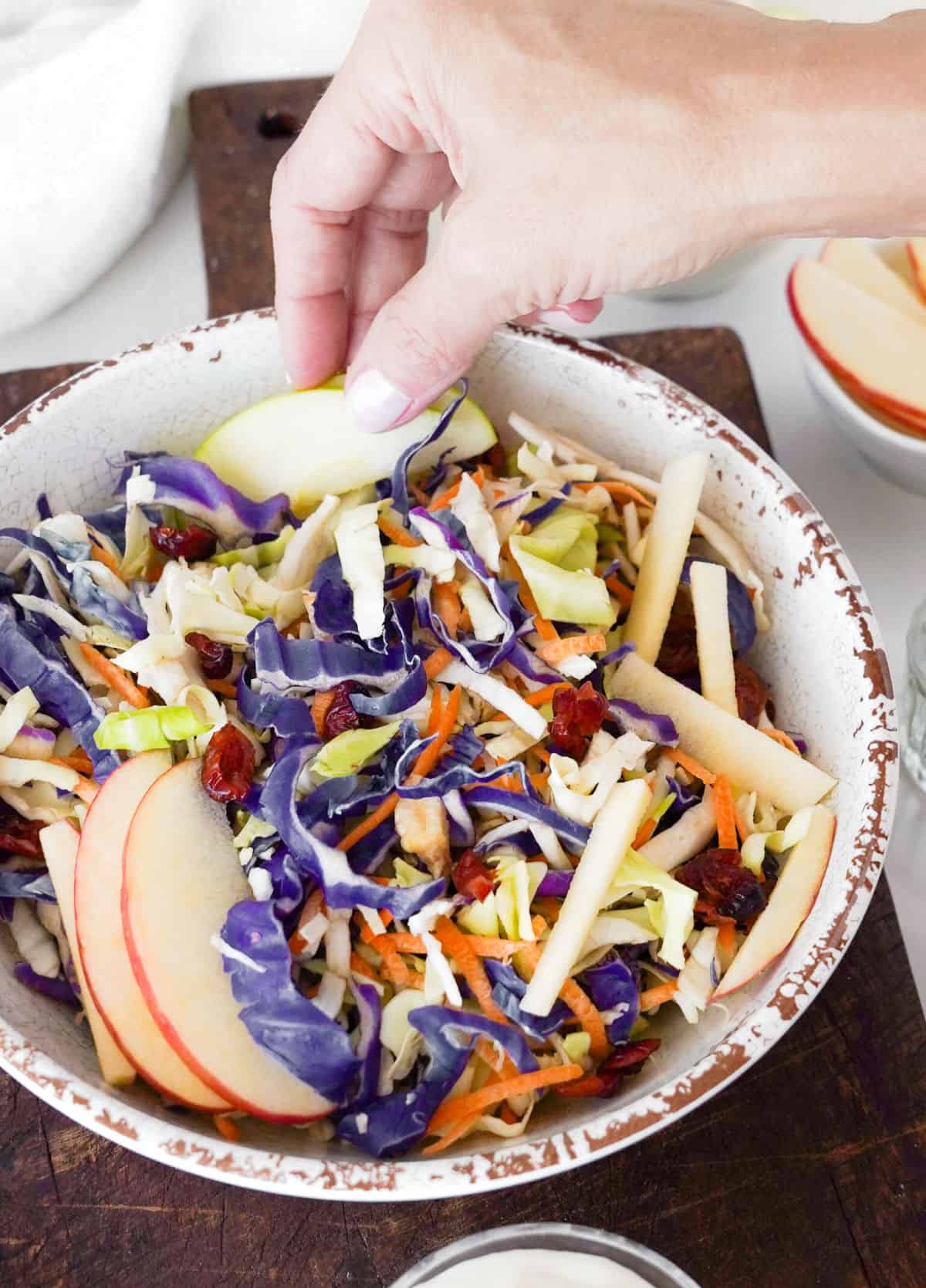 Placing apple slices on a white bowl with coleslaw on a dark wooden board.