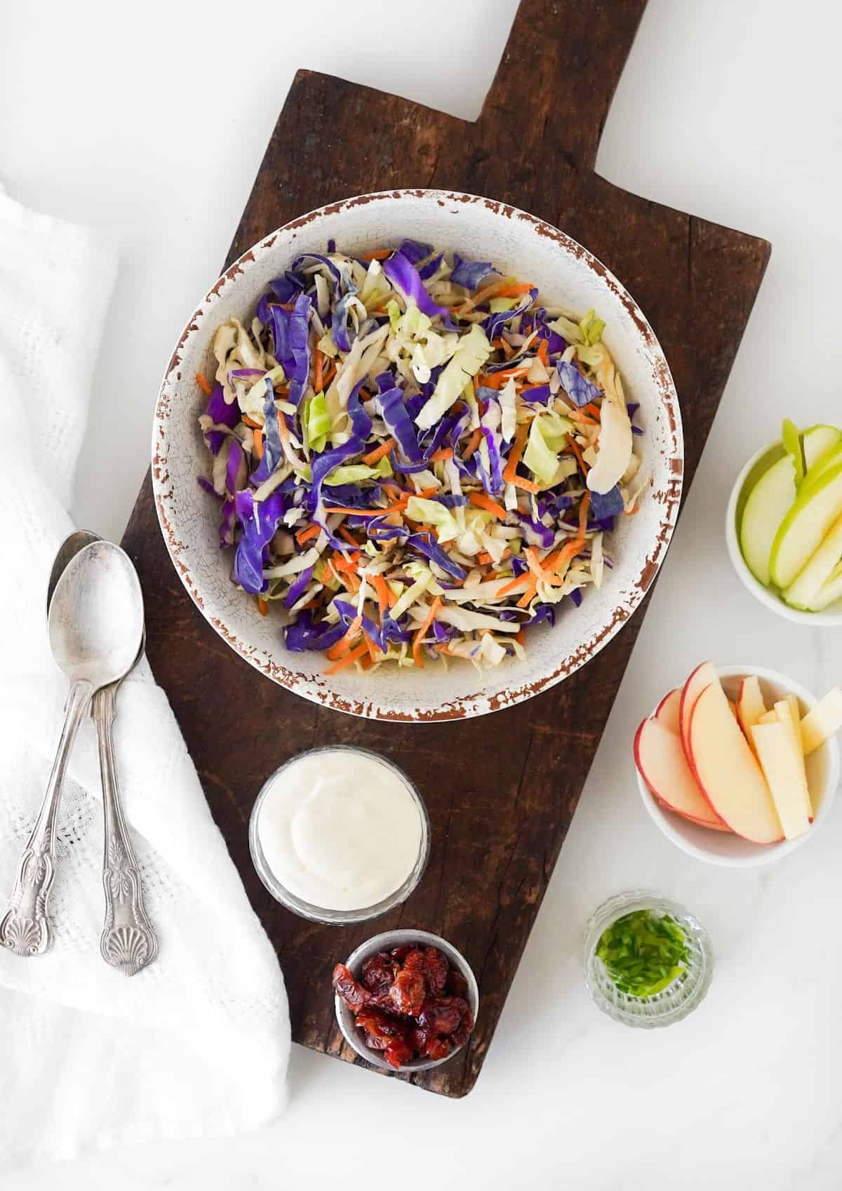 Top view of dark wooden board with bowl of coleslaw, apple slices, dressing, cranberries. Silverware beside it.