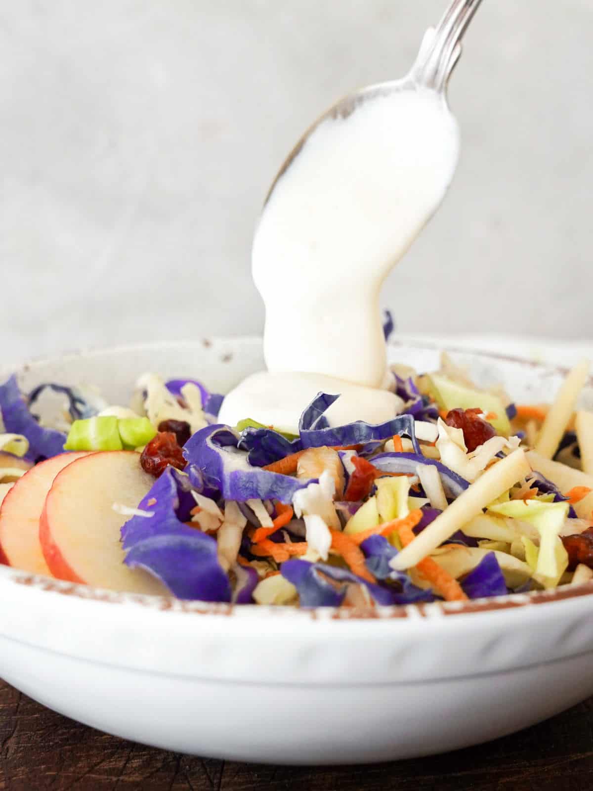 Pouring yogurt dressing with a spoon on a white bowl of cranberry apple coleslaw.