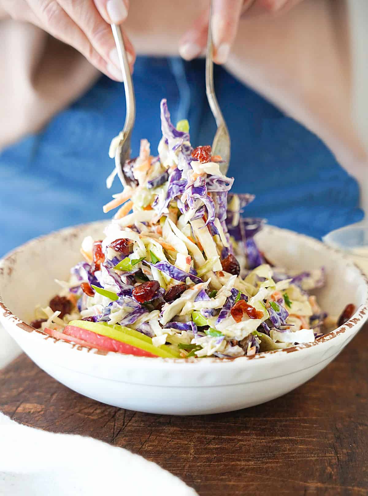 Lifting portion of apple slaw with silver fork and spoon from a white bowl. Wooden surface, jean and sweater background.