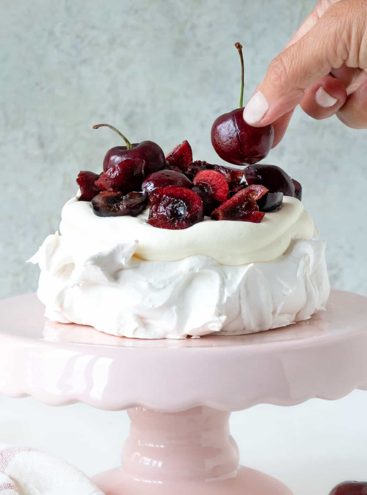 Adding cherries to a cream topped pavlova on a pink cake stand. Grey background. 