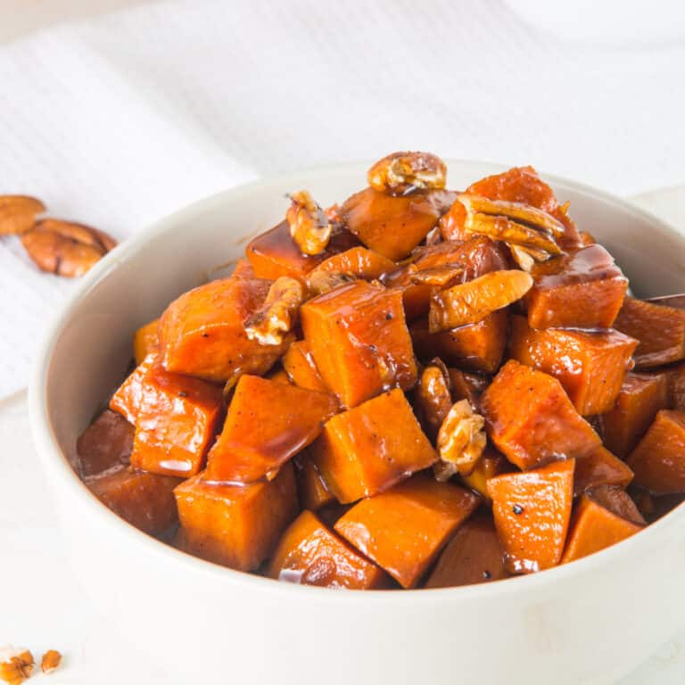 Partial view of white bowl of candied sweet potatoes with pecans on a white surface.