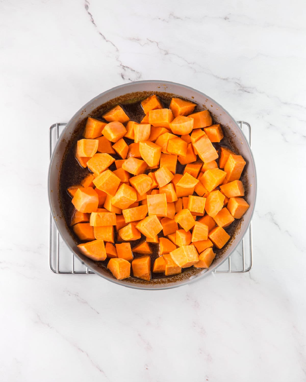 Cubed sweet potatoes in a round handless skillet on a wire rack. White marbled surface.