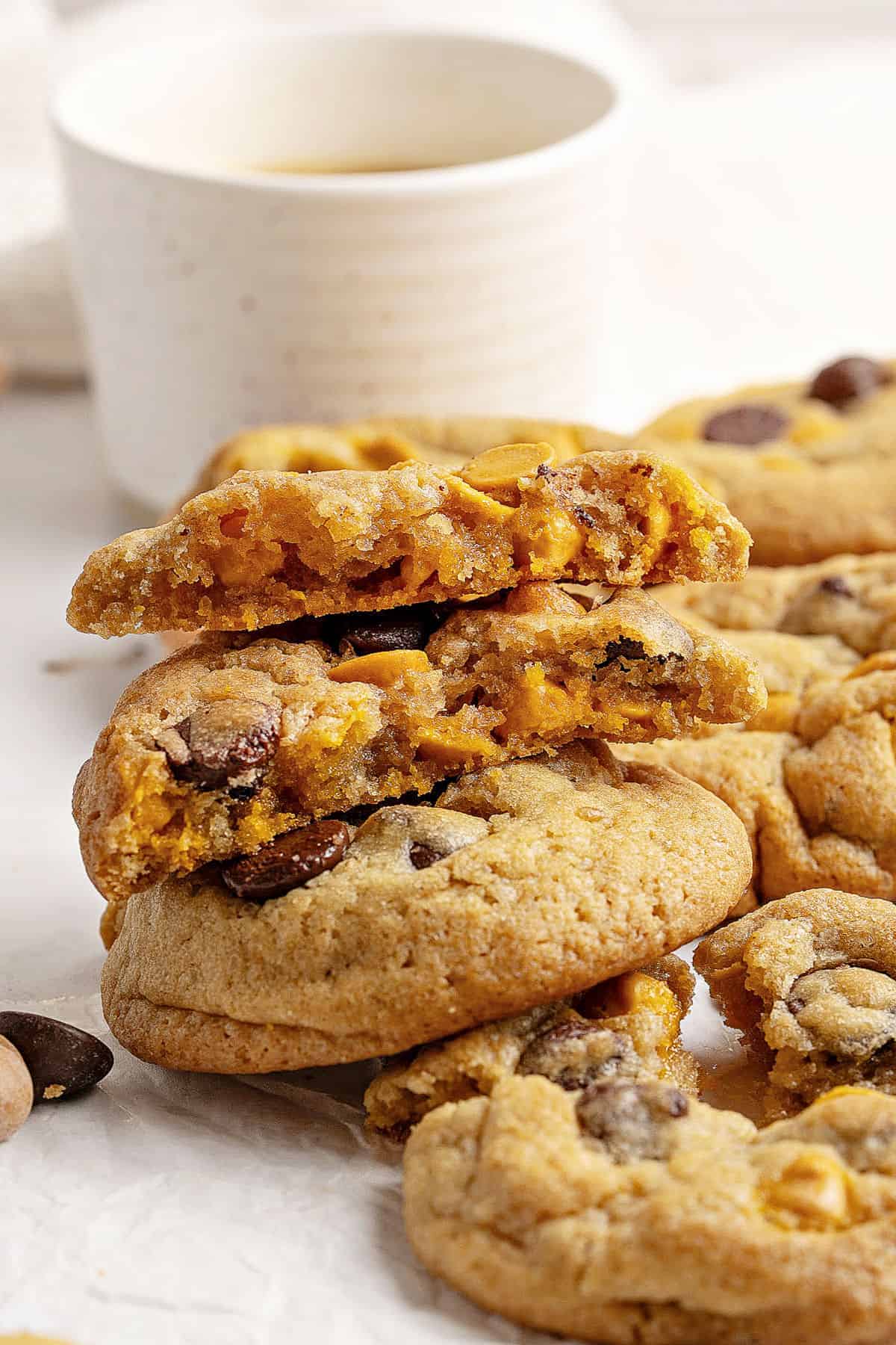 Bitten chocolate chip butterscotch cookies on top of whole ones. Ceramic cup, white background.