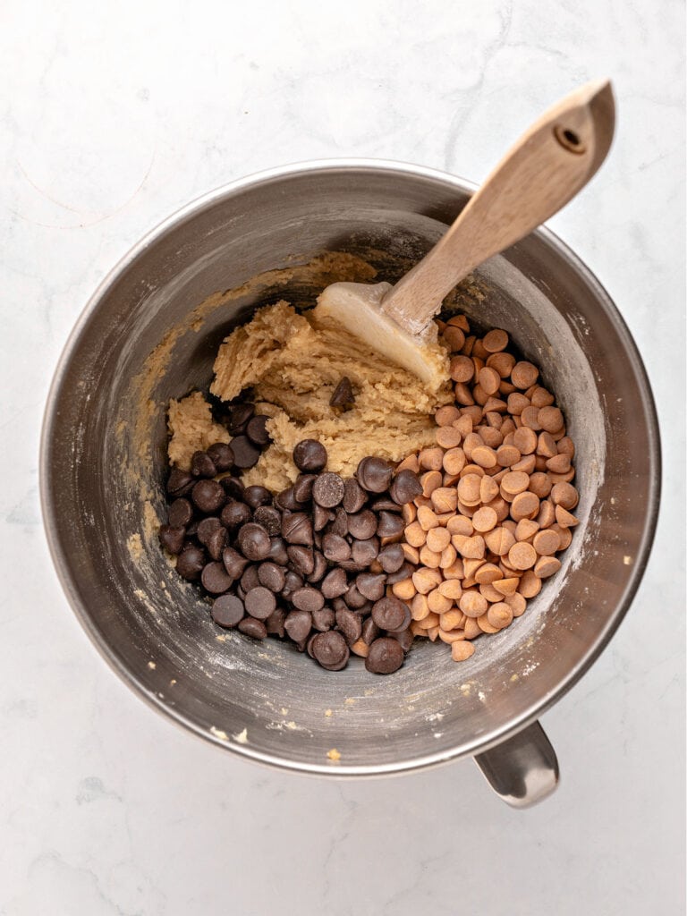 Metal bowl with chocolate and butterscotch chips with cookie dough. Wooden spatula. Light gray background.