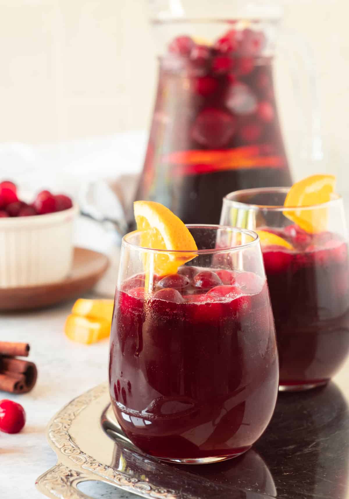 Pitcher and glasses with cranberry sangria and orange pieces. Metal tray, light colored background. 