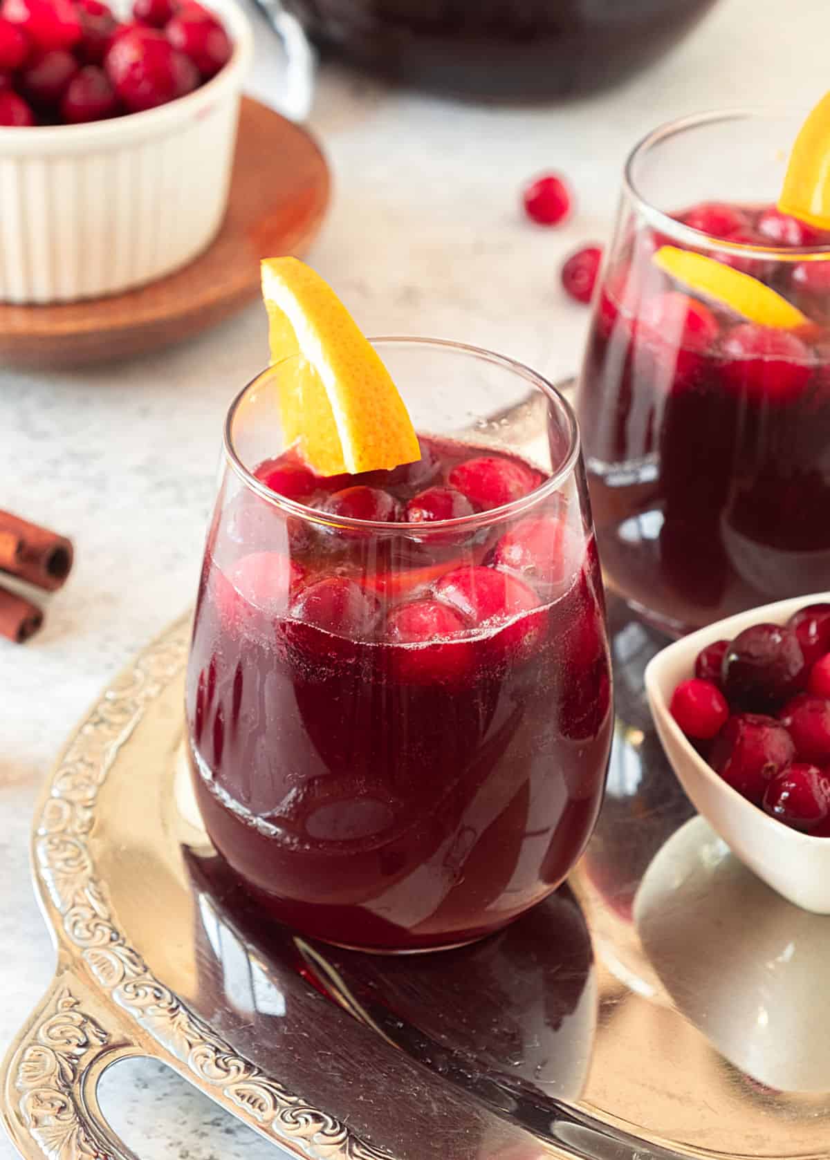 Glasses with cranberry sangria and orange on a metal tray. White bowl with cranberries. Light grey surface. 