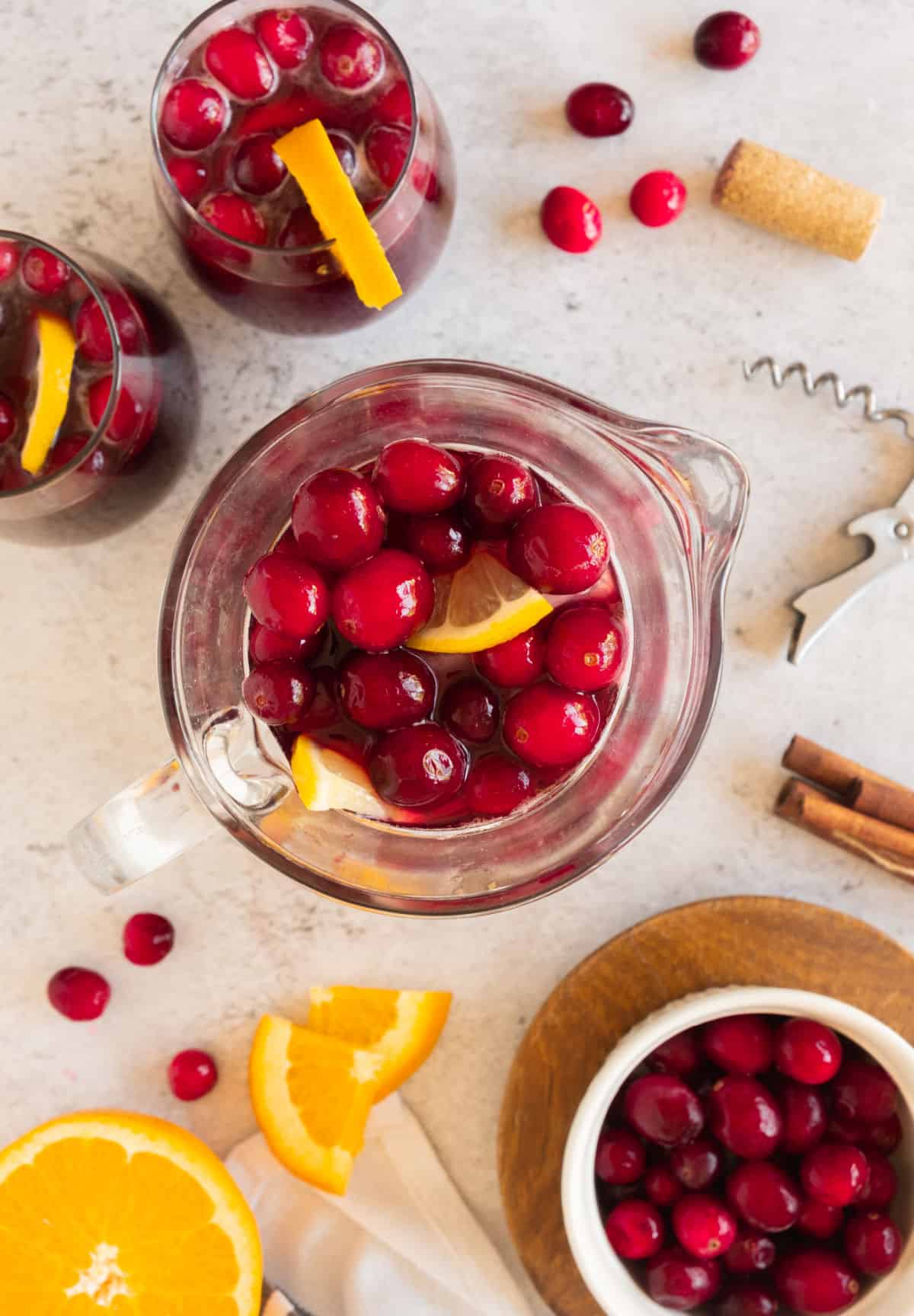 Top view of pitcher with cranberry orange sangria. Light grey surface with orange slices, fresh cranberries, served glasses.