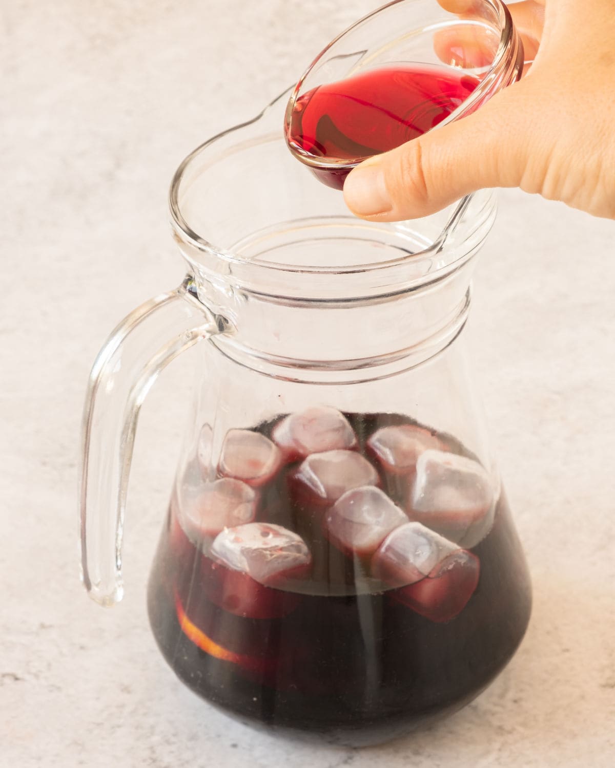 Adding cranberry juice to a pitcher with red wine, citrus slices and ice. Light surface.