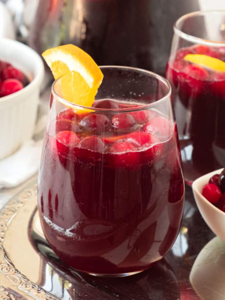 Two glasses of cranberry sangria with orange on a metal tray. White bowls with cranberries.