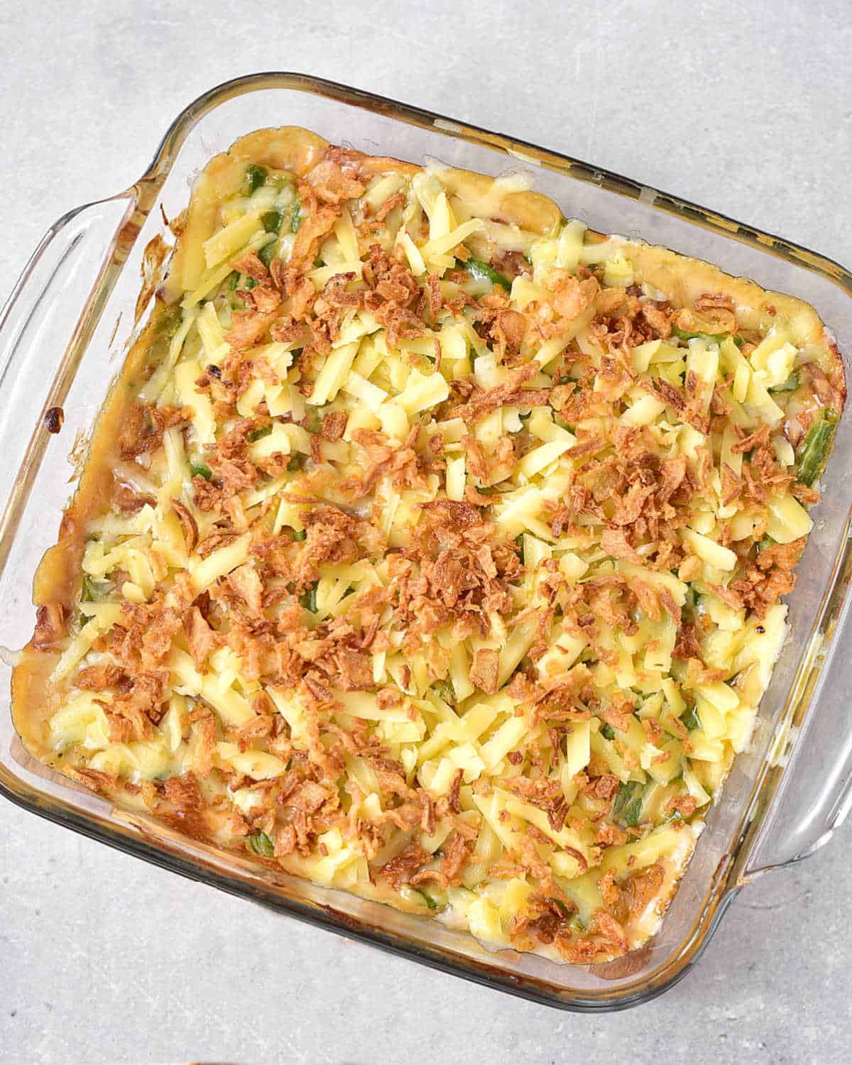 Onion and cheese topped green bean casserole in a glass dish before baking. Grey surface. 