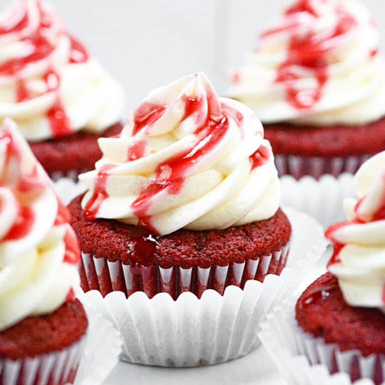 Several frosted red velvet cupcakes in white paper liner with red sauce drizzle. White surface and background.