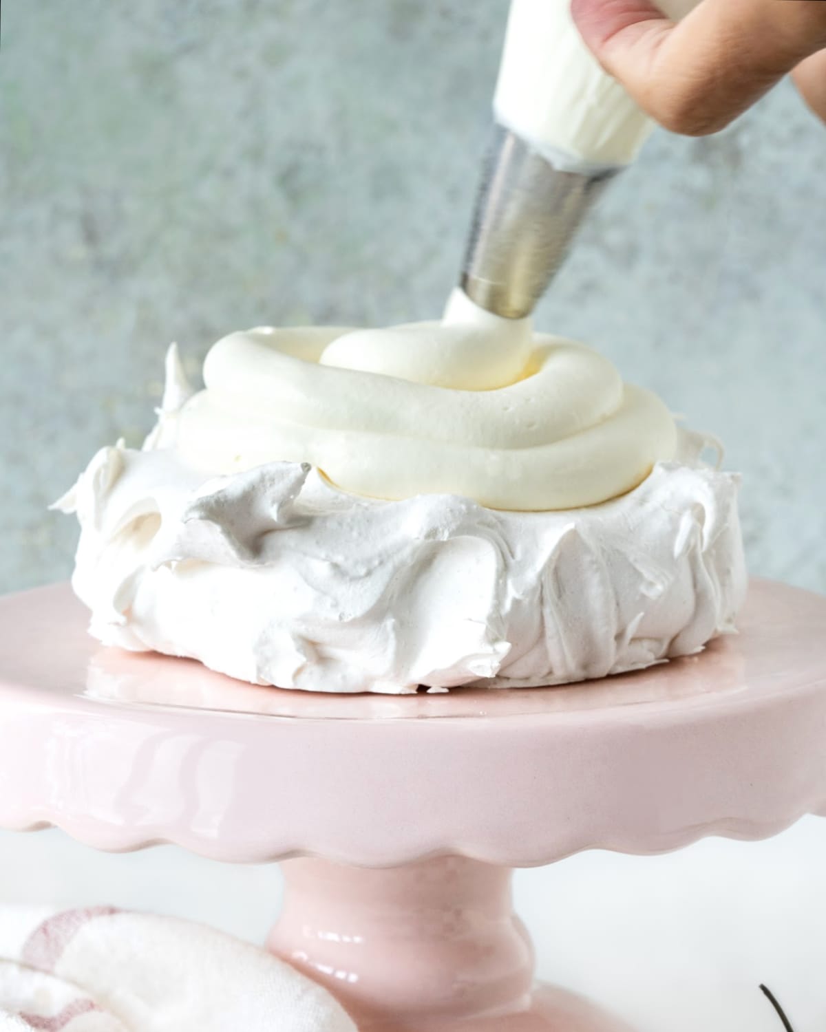 Piping whipped cream on a disc of meringue on a pink cake server. Grey background.