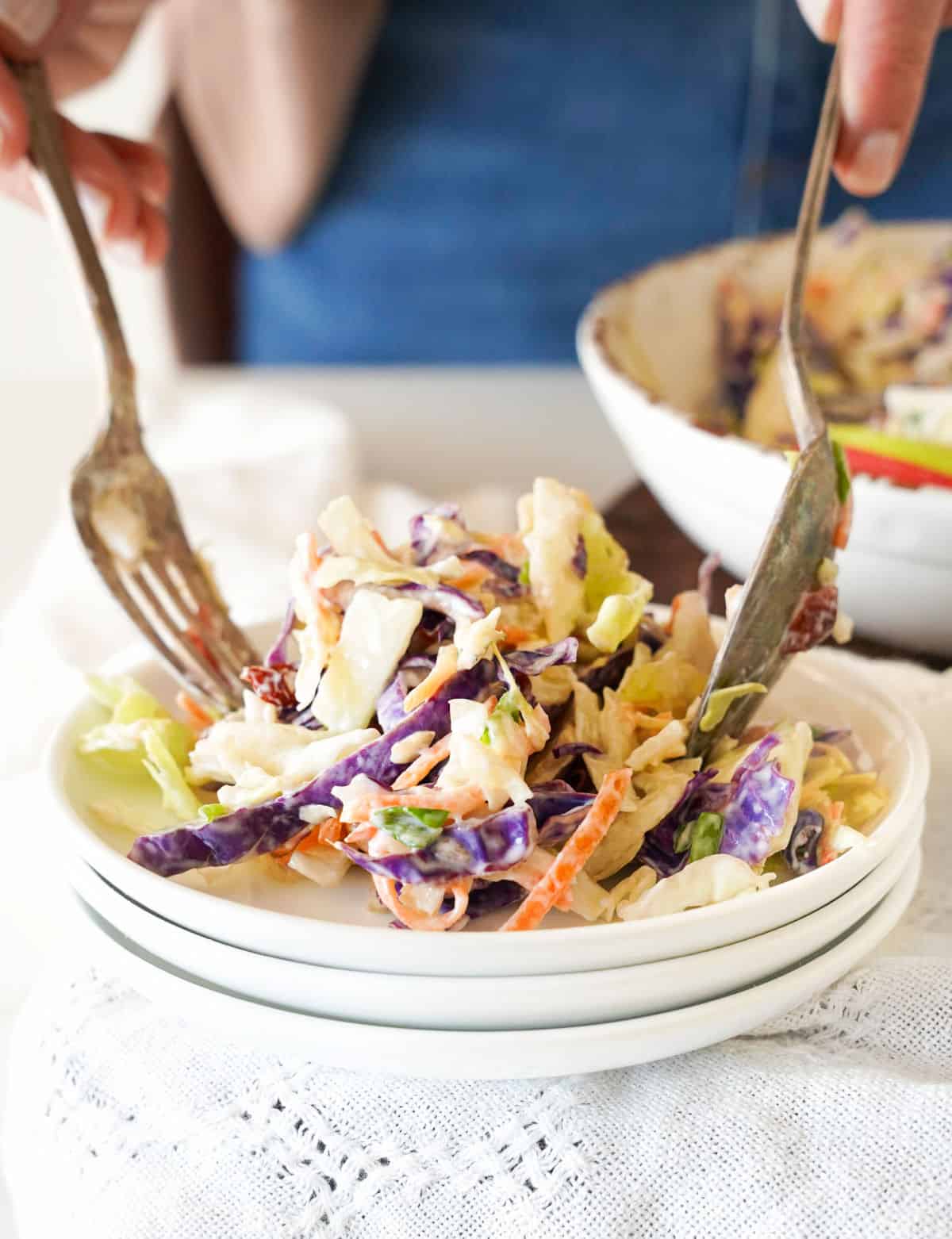 Serving apple coleslaw on a stack of three white plates with silverware. White linen surface.