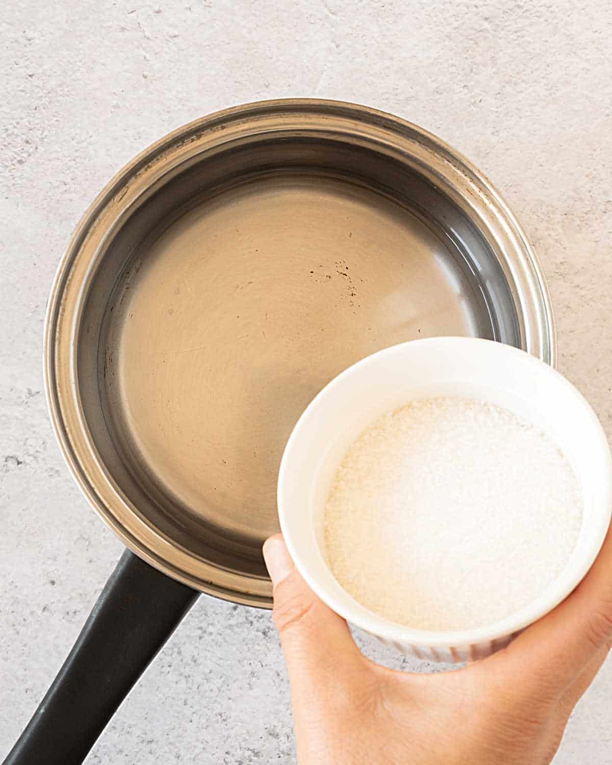 Top view of metal saucepan with water and hand about to pour sugar in a white bowl. Greyish surface.