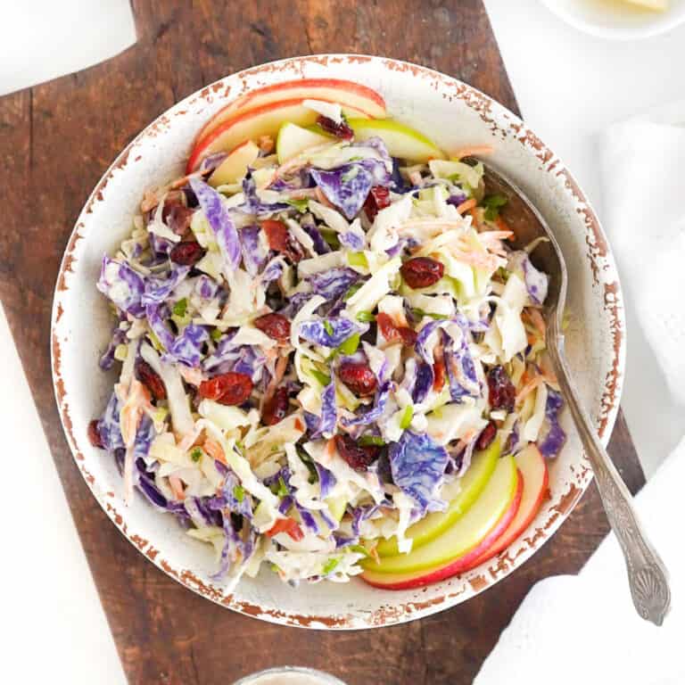 White bowl with silver spoon and cranberry apple slaw on a wooden board.