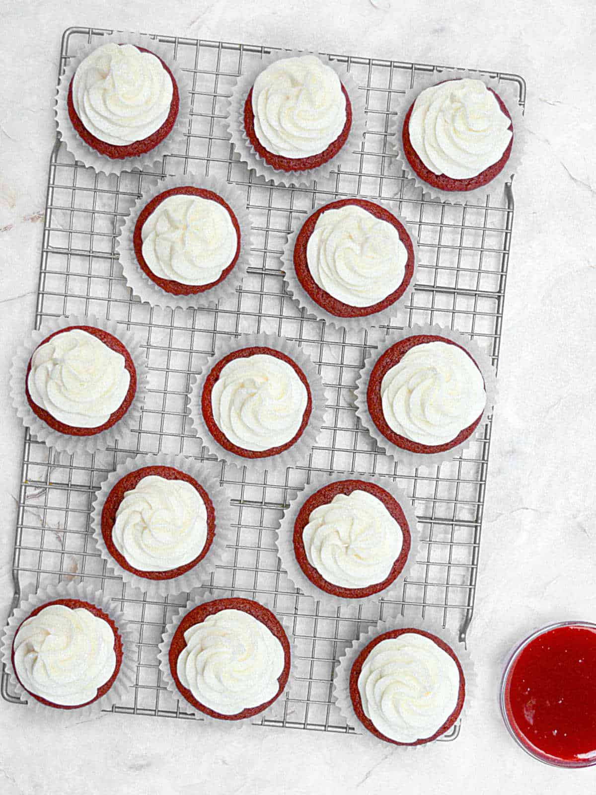 Frosted red velvet cupcakes on a wire rack on a white marbled surface. Bowl with raspberry sauce.
